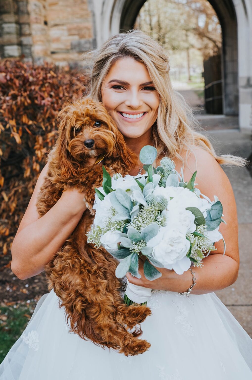 Elopement Wedding A smiling bride in a white wedding dress holds a small, fluffy brown dog in her left arm and a bouquet of white flowers and greenery in her right hand. They stand outdoors in front of a stone archway with autumn foliage in the background. Elopements Inc